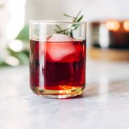 A clear cocktail glass with red liquid, a large ice cube, and a sprig of rosemary.